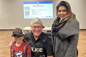 Candidate for Town of Aurora Councillor, Ward 4, Shaheen Moledina (right) smiling with her son (left) and a uniformed police officer (center) in a community meeting hall. They are participating in a local presentation, as indicated by the background projector screen which reads 'Crime Prevention,' demonstrating Moledina's commitment to community safety and family engagement.