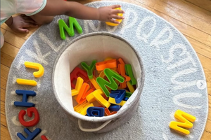 A top-down perspective of a child's hand reaching for a yellow plastic letter on a circular grey alphabet rug at the Active Minds Activity Centre. A fabric bin sits in the center of the rug, overflowing with colorful alphabet toys. This image highlights Shaheen Moledina's professional role as the manager of the centre, where she applies results-oriented leadership to support local families and the educational development of children in Aurora.